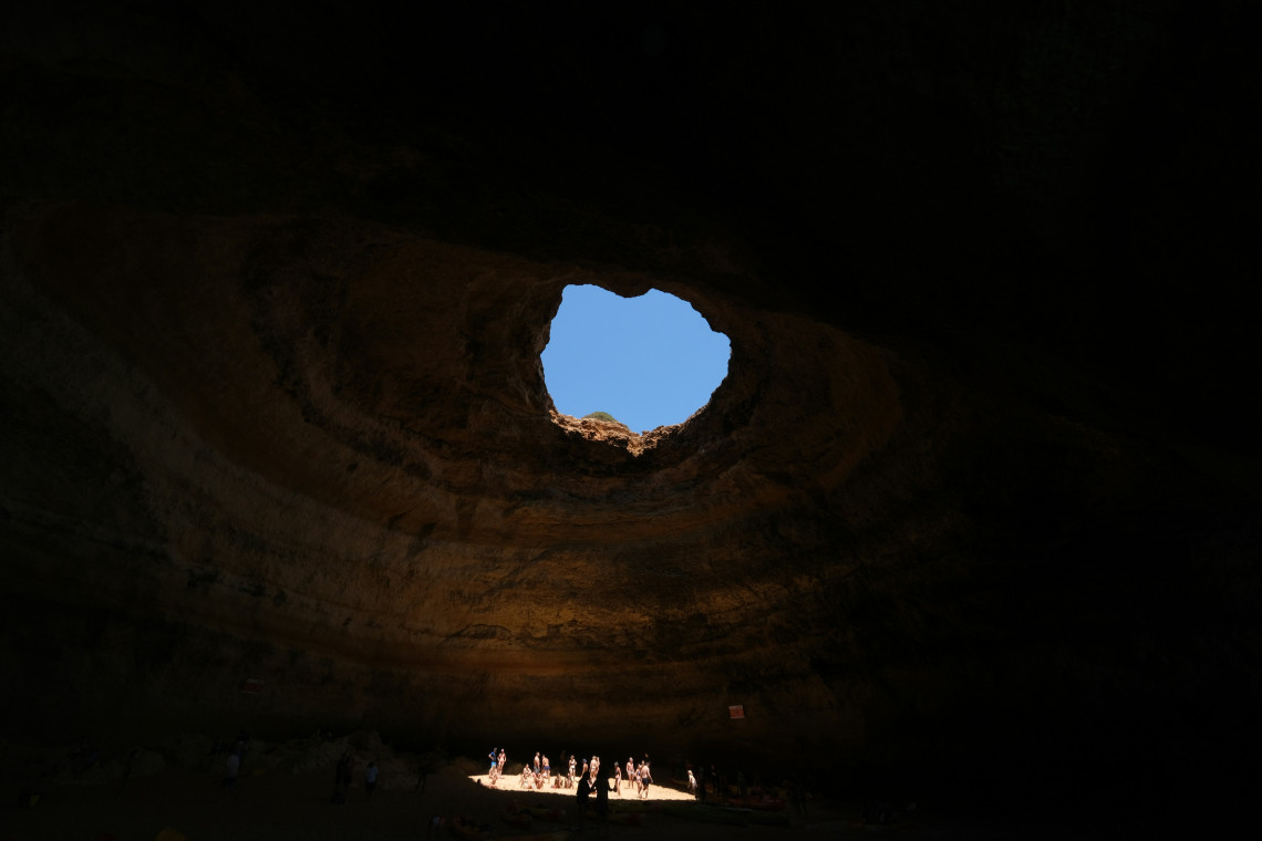View from inside Benagil Sea Cave in Lagoa, Algarve, Portugal, looking up through the circular opening in its limestone ceiling. Sunlight enters the cavern and illuminates visitors gathered on the sandy floor.