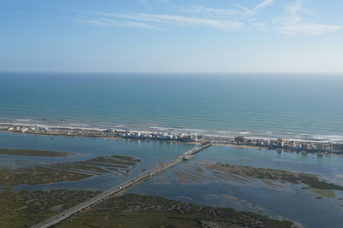 Aerial view of the Ria Formosa lagoon near Faro in the Algarve, Portugal, showing wetlands, channels and the bridge leading toward Faro Island. The Atlantic Ocean and a long line of coastal buildings extend beyond the barrier island.