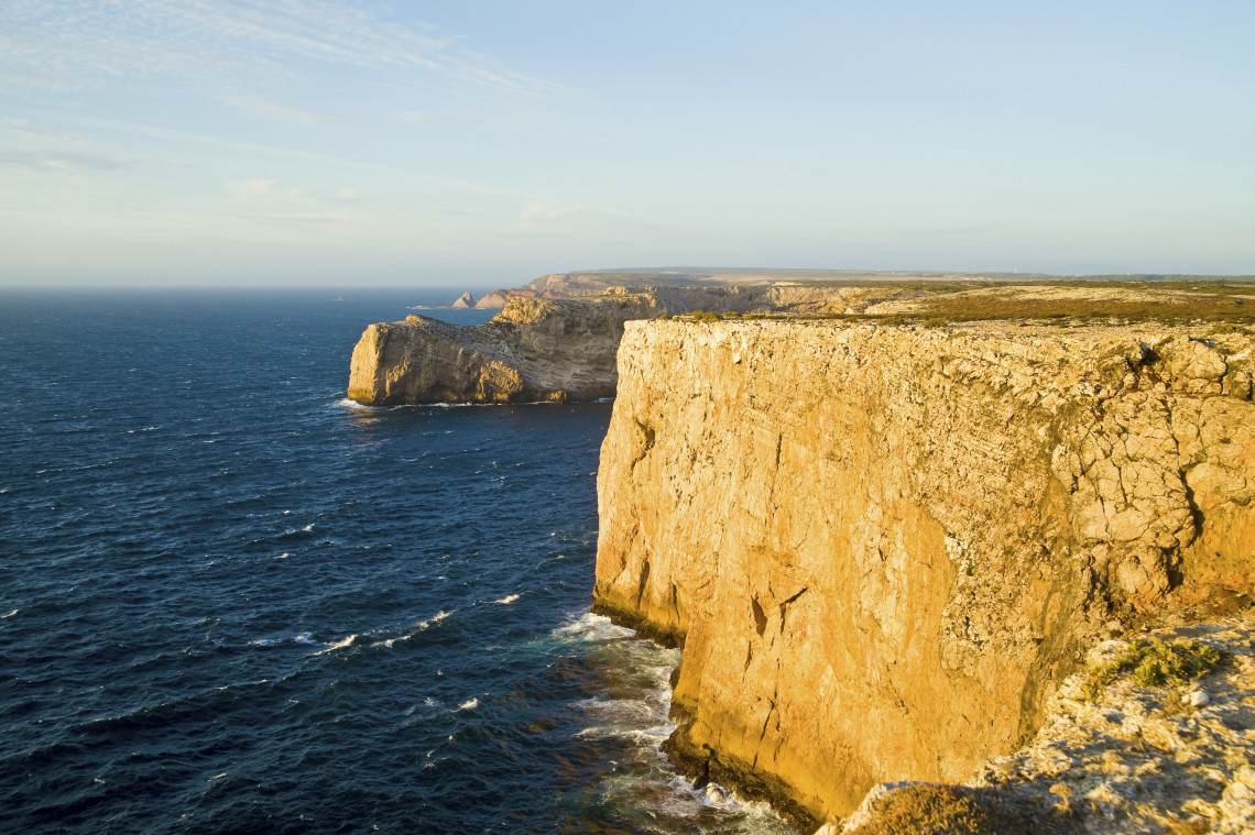 High limestone cliffs meet the deep blue Atlantic Ocean at Cape St Vincent near Sagres in the Algarve, Portugal. Waves break against the base of the exposed headland in warm evening light.