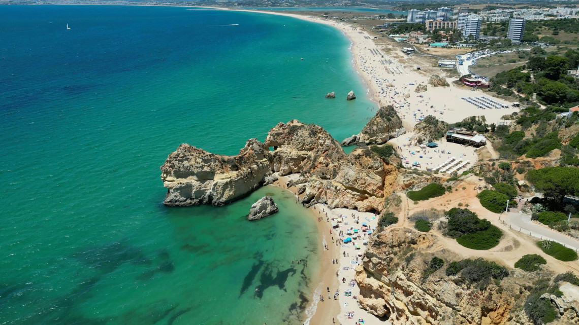 Aerial view of Praia dos Tr&ecirc;s Castelos in Portim&atilde;o, Algarve, Portugal, showing sandy coves, limestone formations and clear turquoise water. The coastline continues toward Praia da Rocha and the city&rsquo;s beachfront buildings.