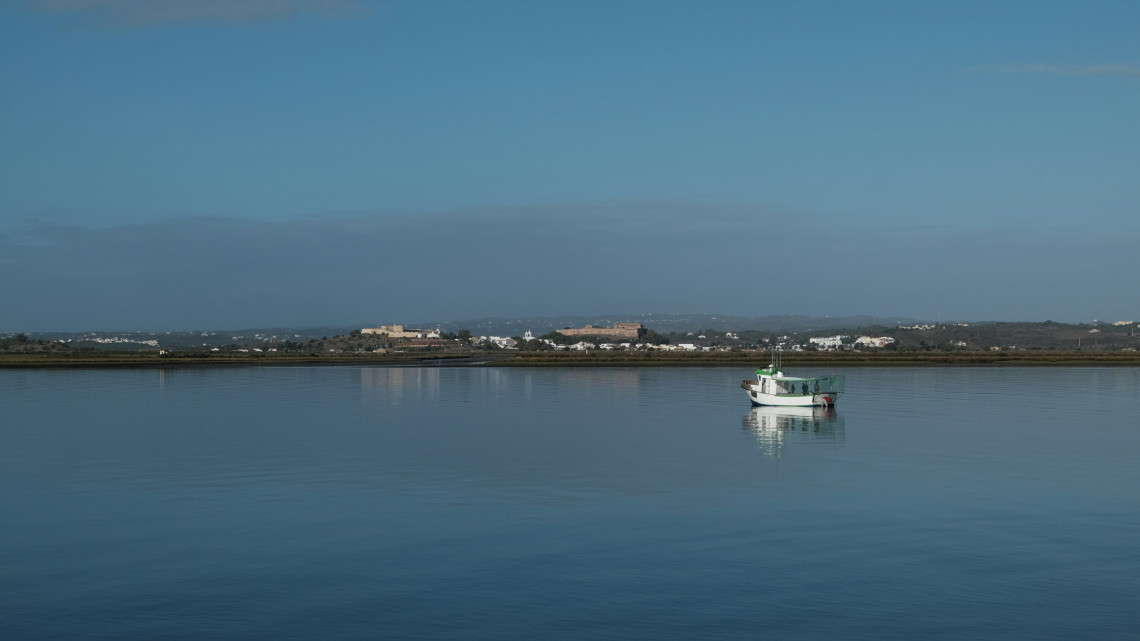 A small fishing boat floats on the calm waters of Ria Formosa near Faro in the Algarve, Portugal. The lagoon reflects the blue sky, with low coastal settlements and distant hills visible along the horizon.
