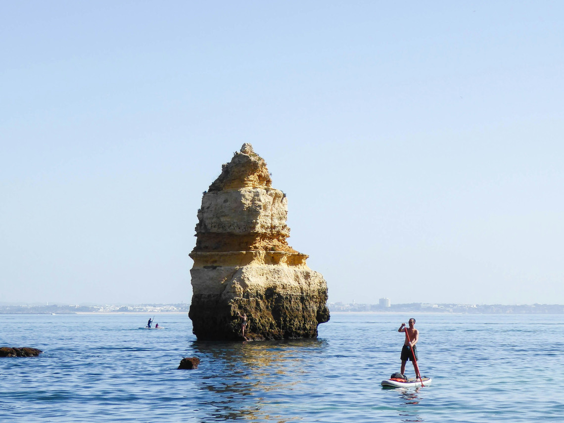 A stand-up paddleboarder moves across the calm Atlantic waters beside a tall limestone sea stack at Ponta da Piedade in Lagos, Algarve, Portugal. The scene highlights the clear coastal waters and distinctive rock formations of the western Algarve.