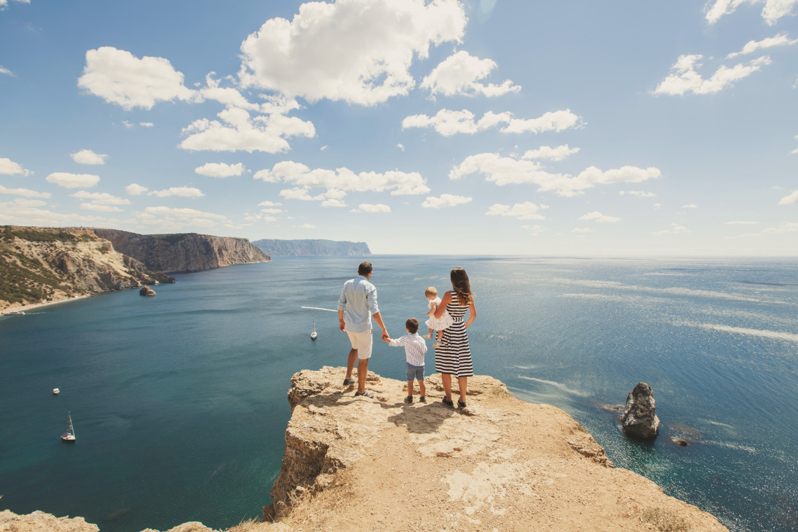 A family stands on a rocky viewpoint above the Atlantic Ocean near Sagres in the Algarve, Portugal. Steep coastal cliffs, offshore rock formations and several sailboats extend across the open seascape.