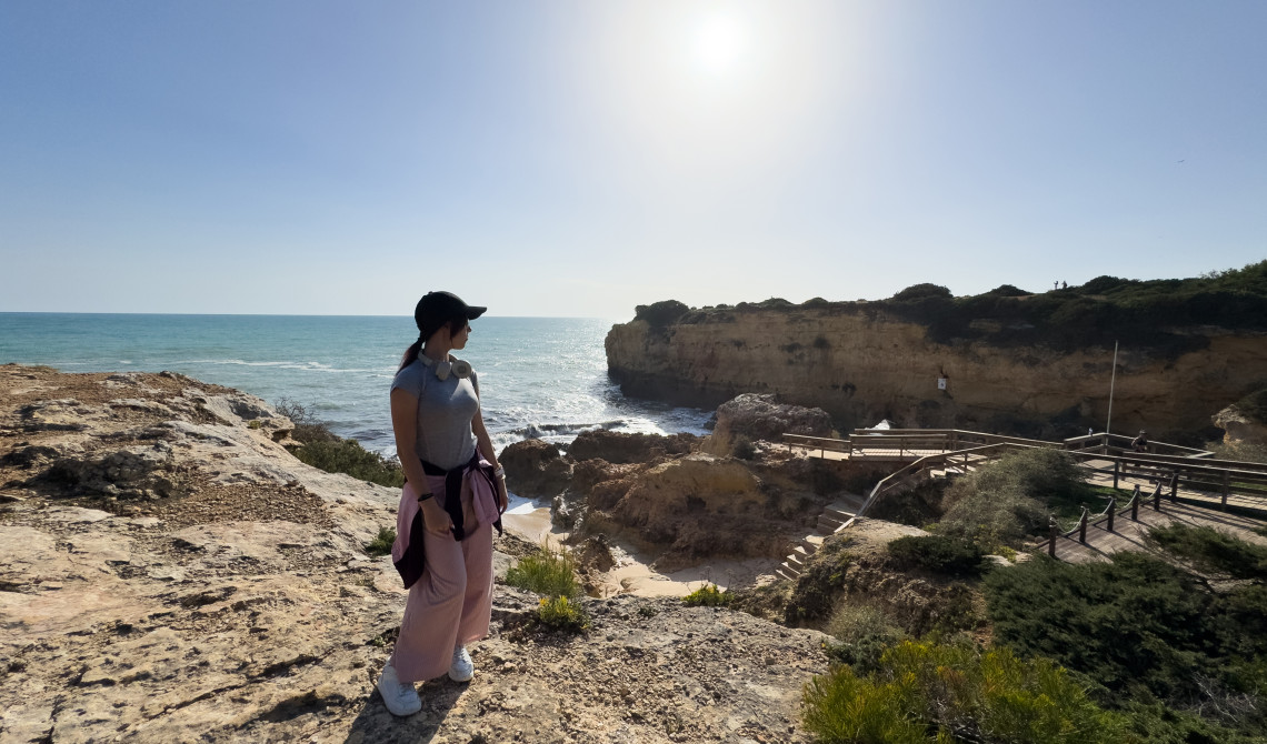 A traveller stands beside the limestone formations and wooden coastal walkway at Algar Seco in Carvoeiro, Algarve, Portugal. The viewpoint overlooks the Atlantic Ocean and the eroded cliffs surrounding the shoreline.