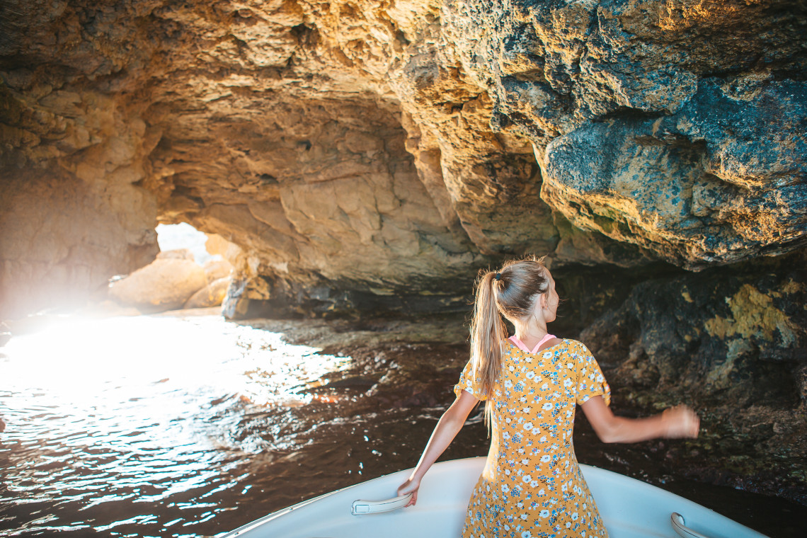 A traveller stands at the front of a boat inside Benagil Sea Cave near Lagoa in the Algarve, Portugal. Sunlight enters through the cave opening and reflects across the water beneath the textured limestone ceiling.