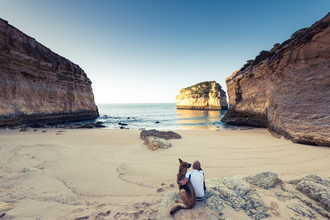 A traveler sits with a dog on the sandy shore of Praia do Carvalho in Lagoa, Algarve, Portugal. Steep limestone cliffs frame the secluded cove, with a large rock formation rising from the clear Atlantic water.