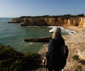 A traveler looks out from the cliffs above Praia da Marinha in Lagoa, Algarve, Portugal. The viewpoint overlooks golden limestone cliffs, a sheltered sandy beach, and the clear waters of the Atlantic Ocean.