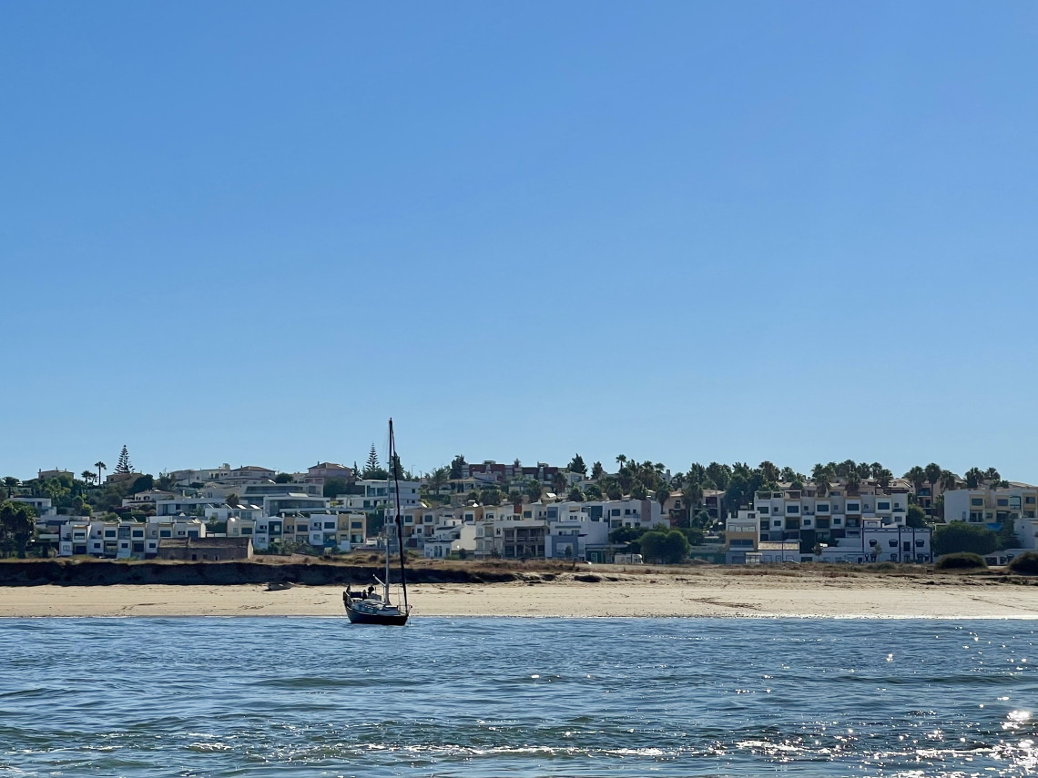 View of Praia de Carvoeiro in the Algarve, Portugal, featuring a sailboat anchored offshore, a sandy beach, and the whitewashed buildings of Carvoeiro rising above the shoreline. The image showcases the traditional charm of one of the Algarve's most picturesque seaside villages.