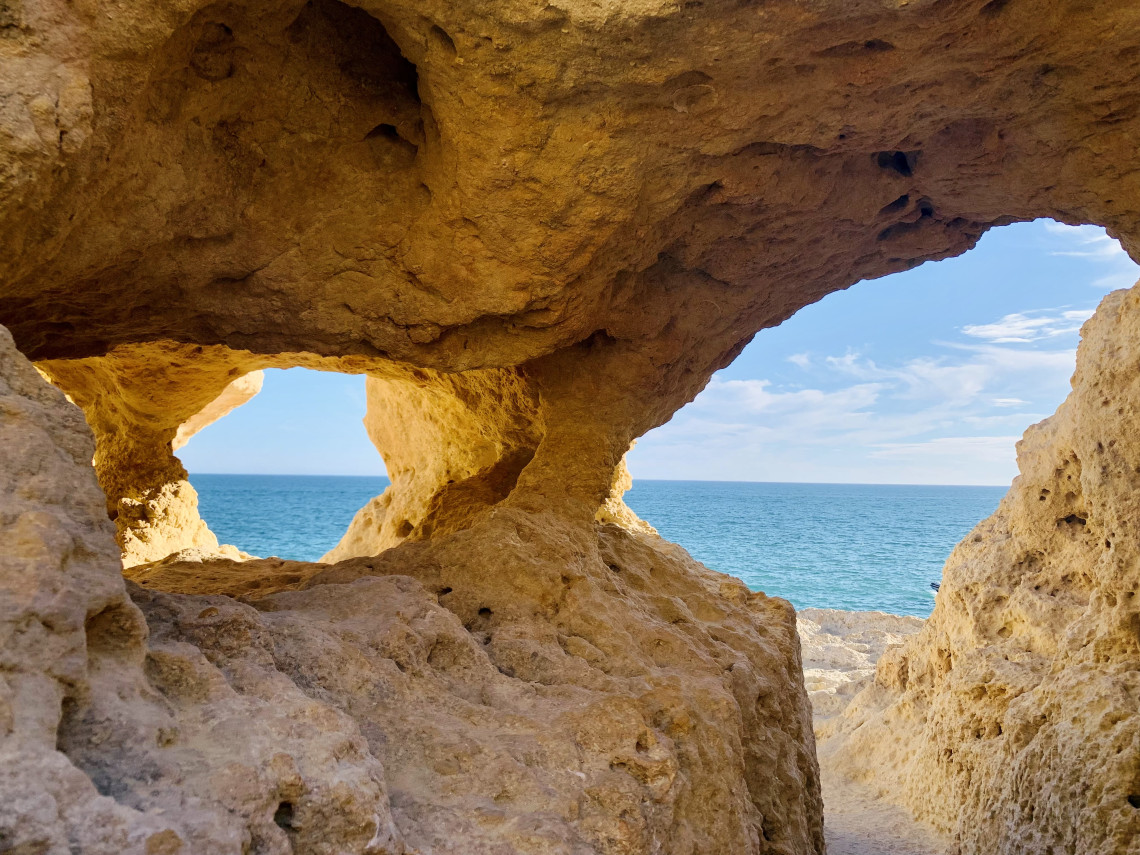 Interior view of a natural sea cave along the Algarve coast in Portugal, featuring weathered limestone rock formations and openings that frame views of the Atlantic Ocean. The image highlights the distinctive geology and coastal landscapes found throughout the Algarve region.