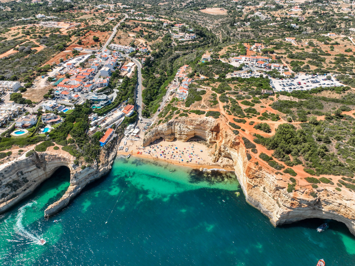 Aerial view of Praia de Benagil in Lagoa, showing the sandy cove, turquoise water, golden limestone cliffs and white buildings extending inland from the coast.