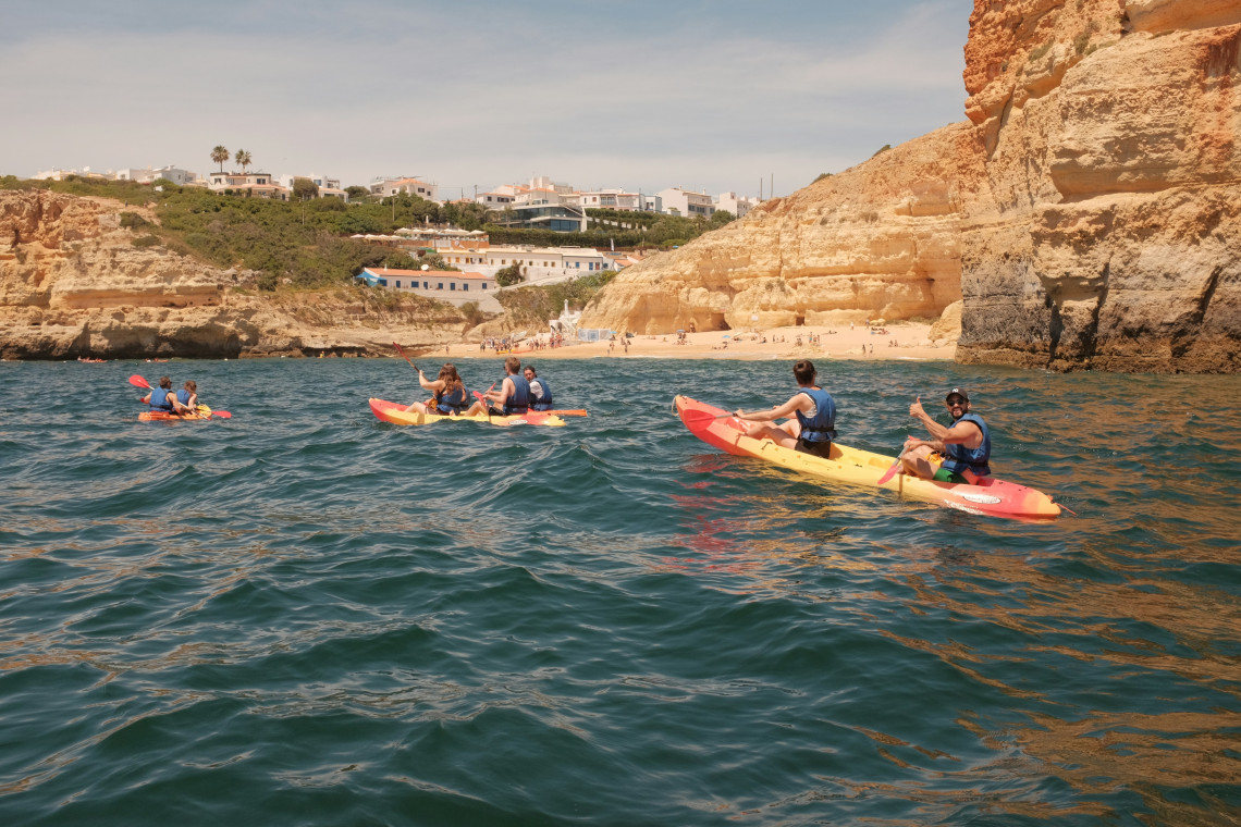 A group of visitors paddles colorful kayaks through the Atlantic waters near Benagil, surrounded by golden limestone cliffs and a sandy cove on the Algarve coast.