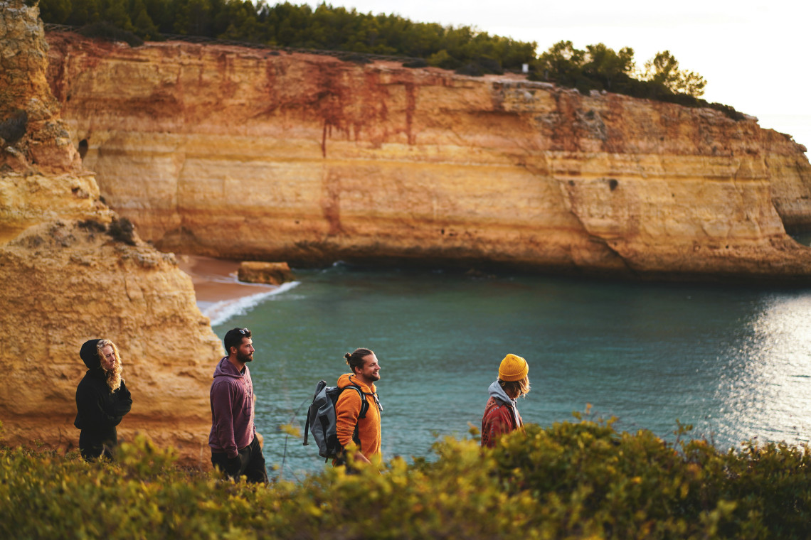 A group of hikers walks along the coastal landscape of Lagoa, with layered golden cliffs, Mediterranean vegetation and a sheltered Atlantic cove in the background.