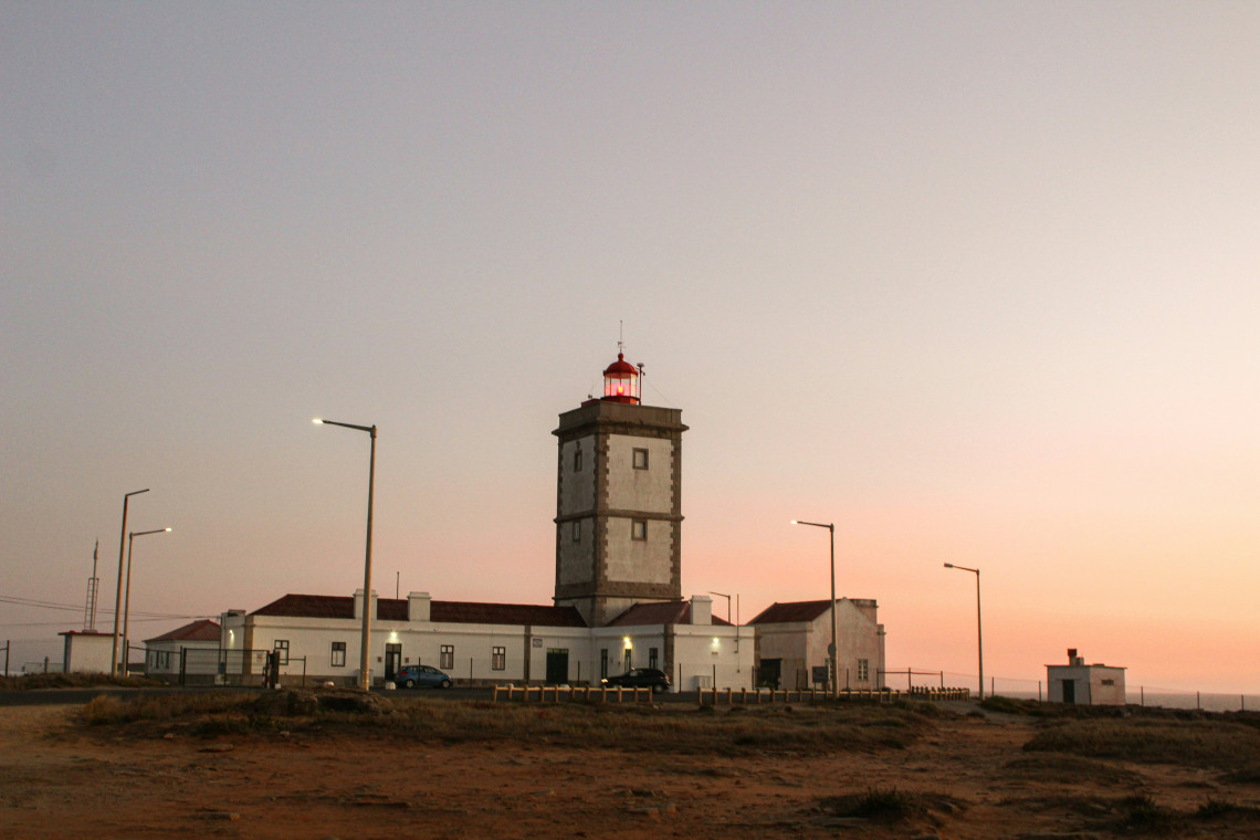 A tall lighthouse with a red lantern stands against a warm sunset sky on the Algarve coast, surrounded by low buildings and rugged coastal terrain.