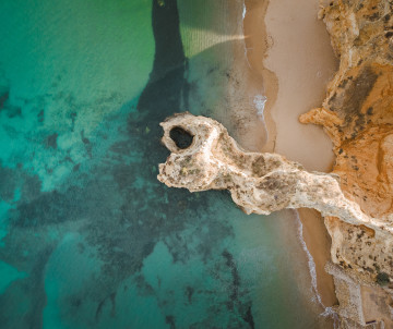 Top-down aerial view of golden limestone formations along the Lagoa coastline, with a natural opening in the rock beside turquoise Atlantic water and a narrow sandy beach.