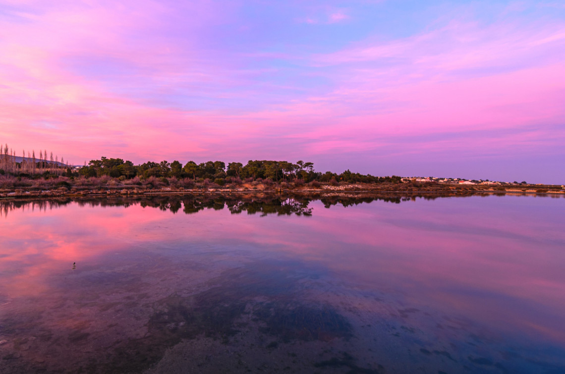 Sunset over Ria Formosa Natural Park in the Algarve, Portugal, with pink and purple skies reflected in the calm waters of the lagoon. The image showcases the protected coastal wetlands, salt marshes, and natural landscapes that make Ria Formosa one of Portugal's most important nature reserves.