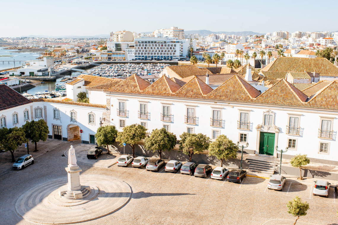 Elevated view of Faro Old Town in Faro, Algarve, Portugal, featuring historic whitewashed buildings, traditional terracotta rooftops, a central square, and the city's marina in the background. The image showcases the blend of historic architecture and waterfront scenery that characterizes the Algarve's capital city.