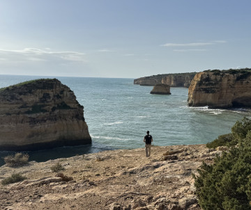 Panoramic view of the Algarve coastline in Portugal featuring limestone cliffs, sea stacks, and the Atlantic Ocean. A visitor stands on a rocky viewpoint overlooking the rugged shoreline and coastal rock formations characteristic of southern Portugal.
