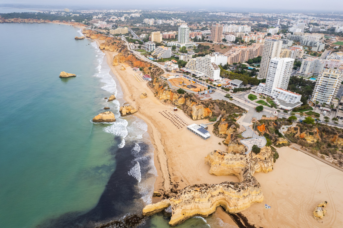 Aerial view of Praia da Rocha in Portim&atilde;o, Algarve, Portugal, featuring golden limestone cliffs, a wide sandy beach, turquoise Atlantic waters, and the city's waterfront skyline. The image showcases one of the Algarve's most famous beach destinations and coastal landscapes.