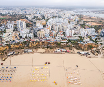 Aerial view of Praia da Rocha in Portimão, Algarve, Portugal, showing the wide sandy beach lined with sun loungers and the city skyline rising behind the coastline. The image highlights one of the Algarve's most famous beach destinations and its vibrant s
