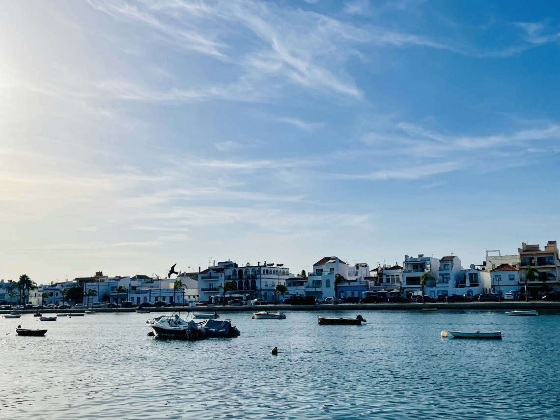 View of the waterfront along the Gil&atilde;o River in Tavira, Algarve, Portugal, with traditional whitewashed buildings and small boats moored on the calm water. The image showcases the historic charm and maritime heritage of one of the Algarve's most picturesque towns.