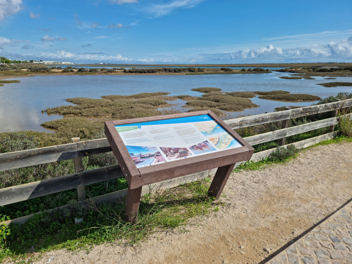 View of the Ria Formosa Natural Park near Tavira, Algarve, Portugal, featuring salt marshes, tidal lagoons, and an interpretive information panel along a walking trail. The protected wetland is one of Portugal's most important coastal ecosystems and a popular destination for birdwatching and nature exploration.