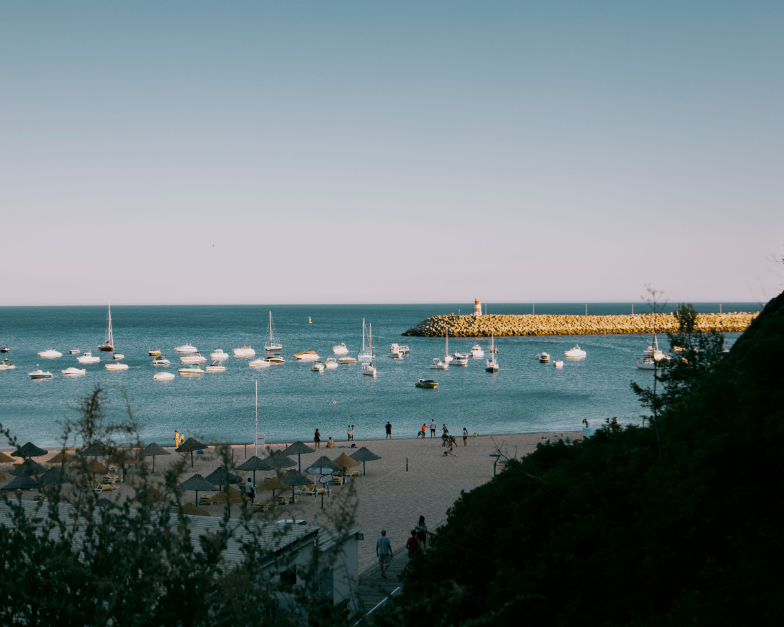 View over a sandy Algarve beach and sheltered marina, with sailboats and small vessels anchored behind a protective breakwater on the Atlantic coast.
