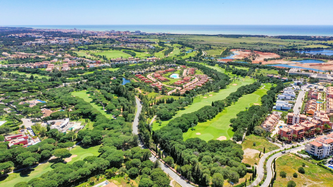 Aerial view across several landscaped golf courses in the Algarve, surrounded by pine trees, resorts, residential areas and the Atlantic coastline in the distance.