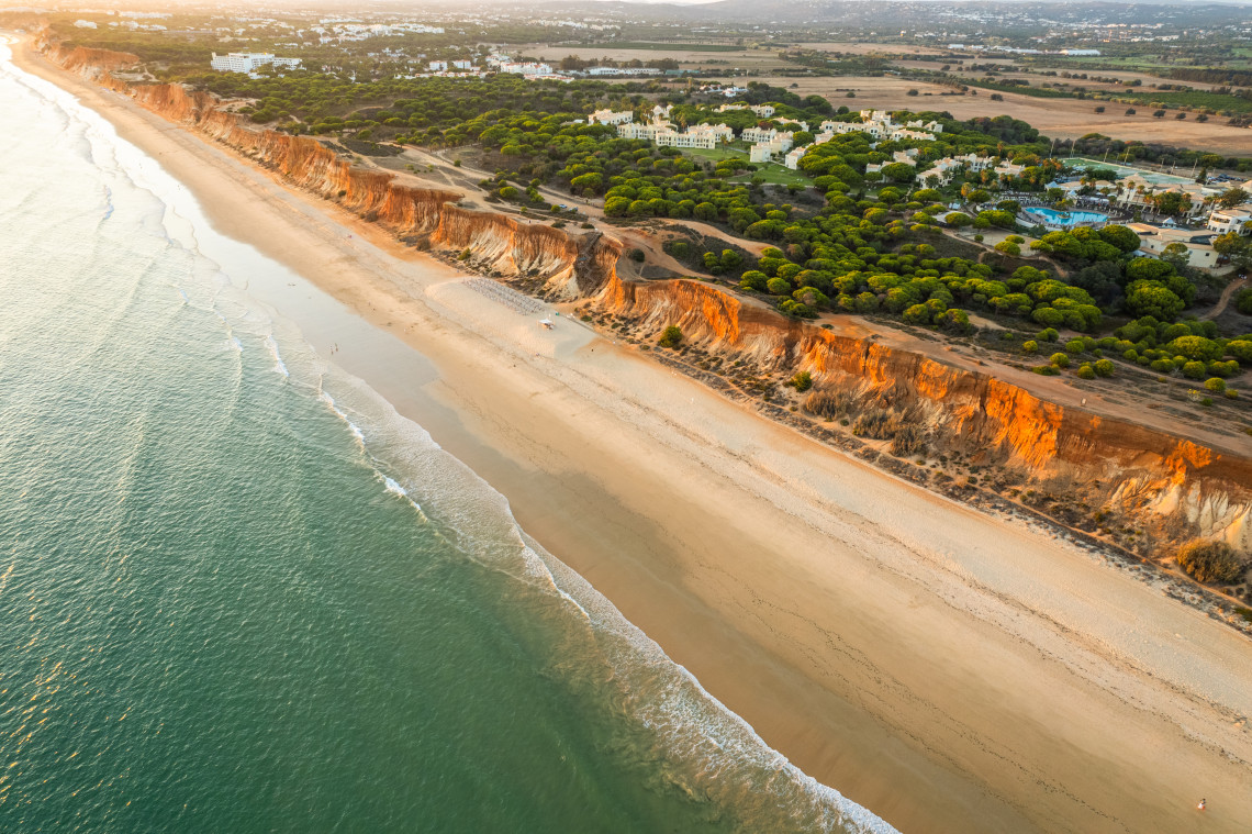 Aerial coastal view of Fal&eacute;sia Beach showing extended white sandy shoreline, distinctive orange-red cliff formations, turquoise Atlantic Ocean, and resort development nestled in green vegetation in the Algarve